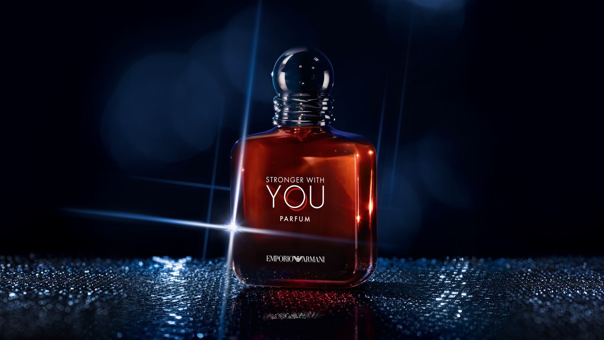  A single bottle of Emporio Armani "Stronger With You Parfum" stands upright on a reflective surface, illuminated by dramatic blue and white lighting against a dark background.