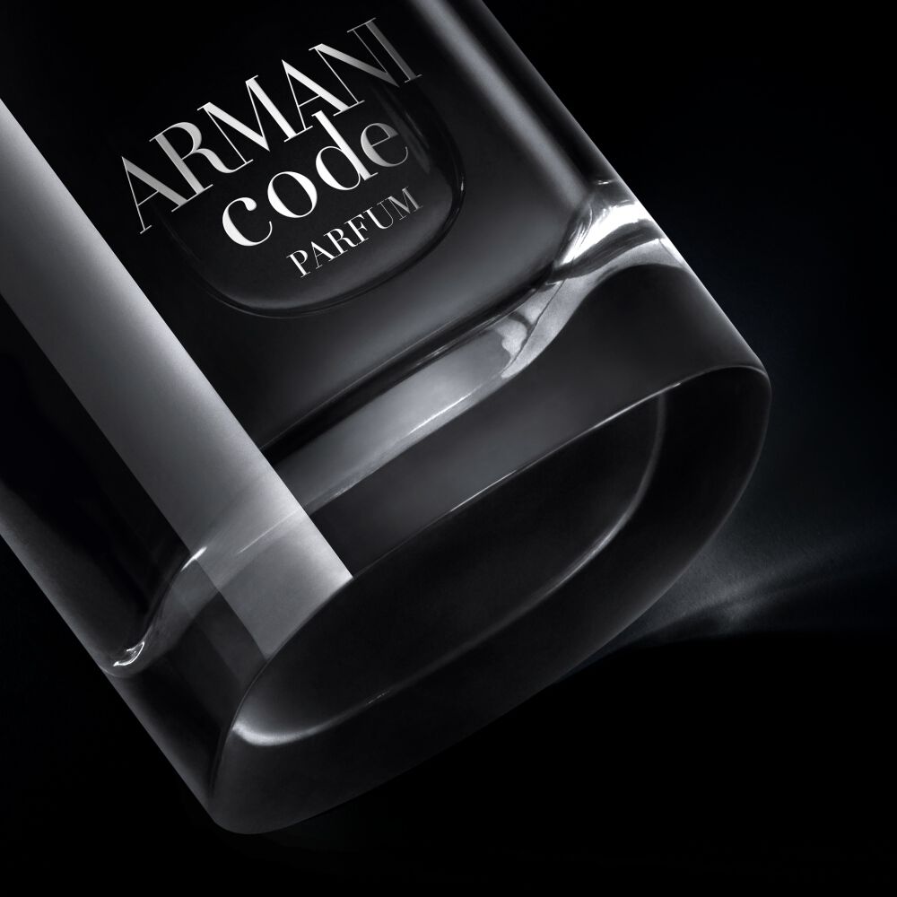A close-up, angled view of the bottom of an Armani Code Parfum bottle, showcasing its clear glass base and part of the dark body with the silver logo, dramatically lit against a black background.