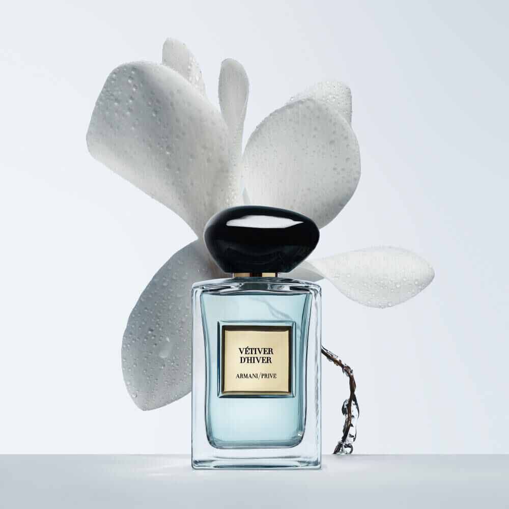 Armani Prive Les Eaux Vetiver d'Hiver eau de parfum, transparent rectangular glass bottle with black rounded cap, beside a sculptural white flower on a pale grey-blue background.
