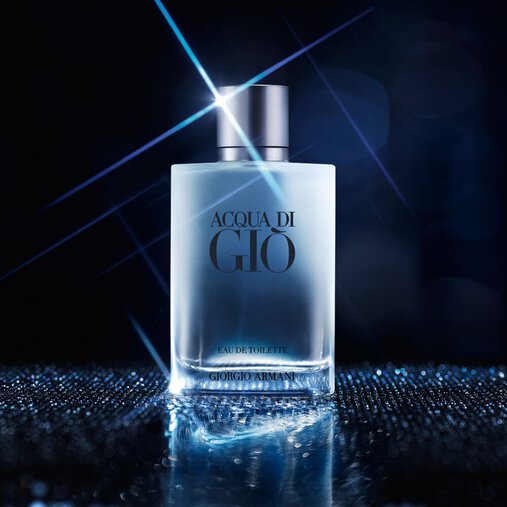 A bottle of Acqua di Gio fragrance with a clear, frosted appearance and a silver cap is centrally placed on a shimmering, reflective surface. A bright starburst light effect emanates from the top of the bottle, and the background is a dark, blurry blue with subtle light bokeh, creating a sophisticated and ethereal atmosphere.