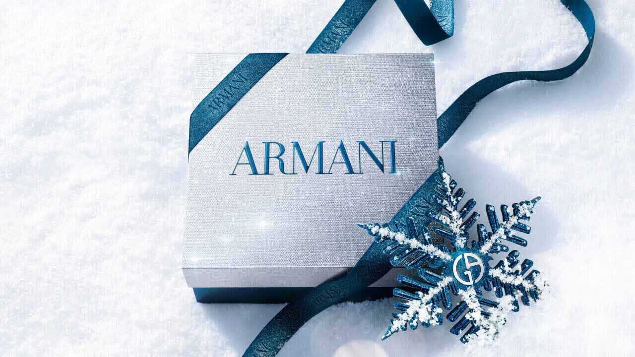 Armani beauty gift box — silver gift box wrapped with blue satin ribbon and a metallic snowflake ornament, resting on fresh white snow.