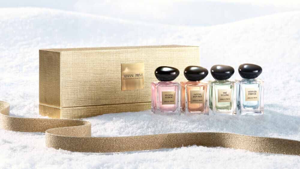Armani Prive Les Eaux Holiday miniature gift set: gold rectangular box alongside four miniature eau de parfum bottles, photographed on a pale textured snow surface.