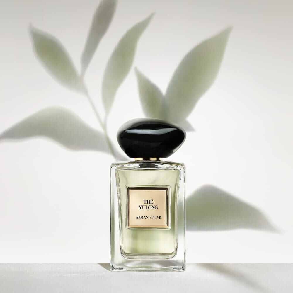 Armani Prive Les Eaux The Yulong eau de parfum, transparent rectangular glass bottle with black rounded cap, set against delicate green botanical leaves on a pale sage background.