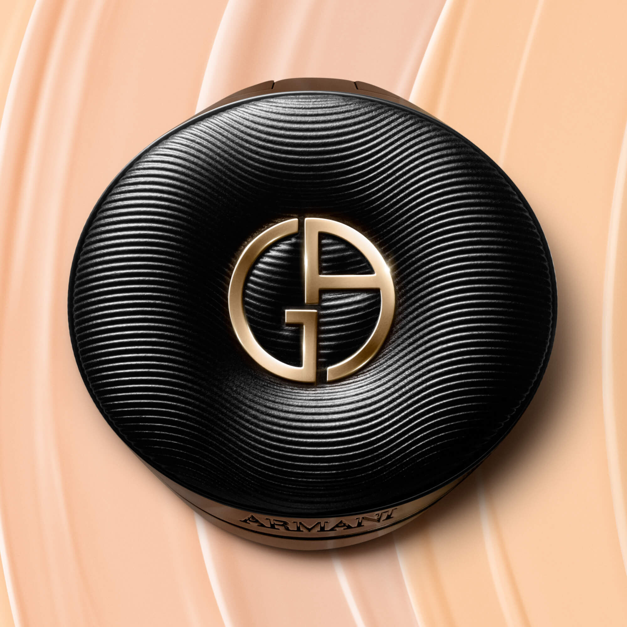 The closed black Giorgio Armani LUMINOUS SILK PRIMA GLOW CUSHION compact, featuring a textured lid and a golden GA logo, set against a background of wavy, light beige foundation strokes.