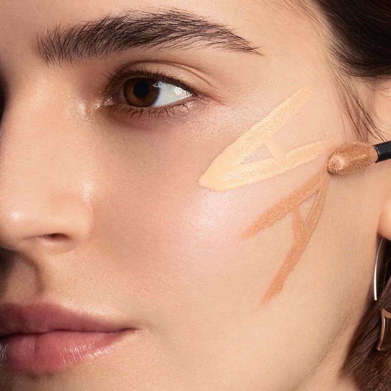 A model's face with two shades of liquid foundation or concealer applied in V-shapes on their cheek and jawline.