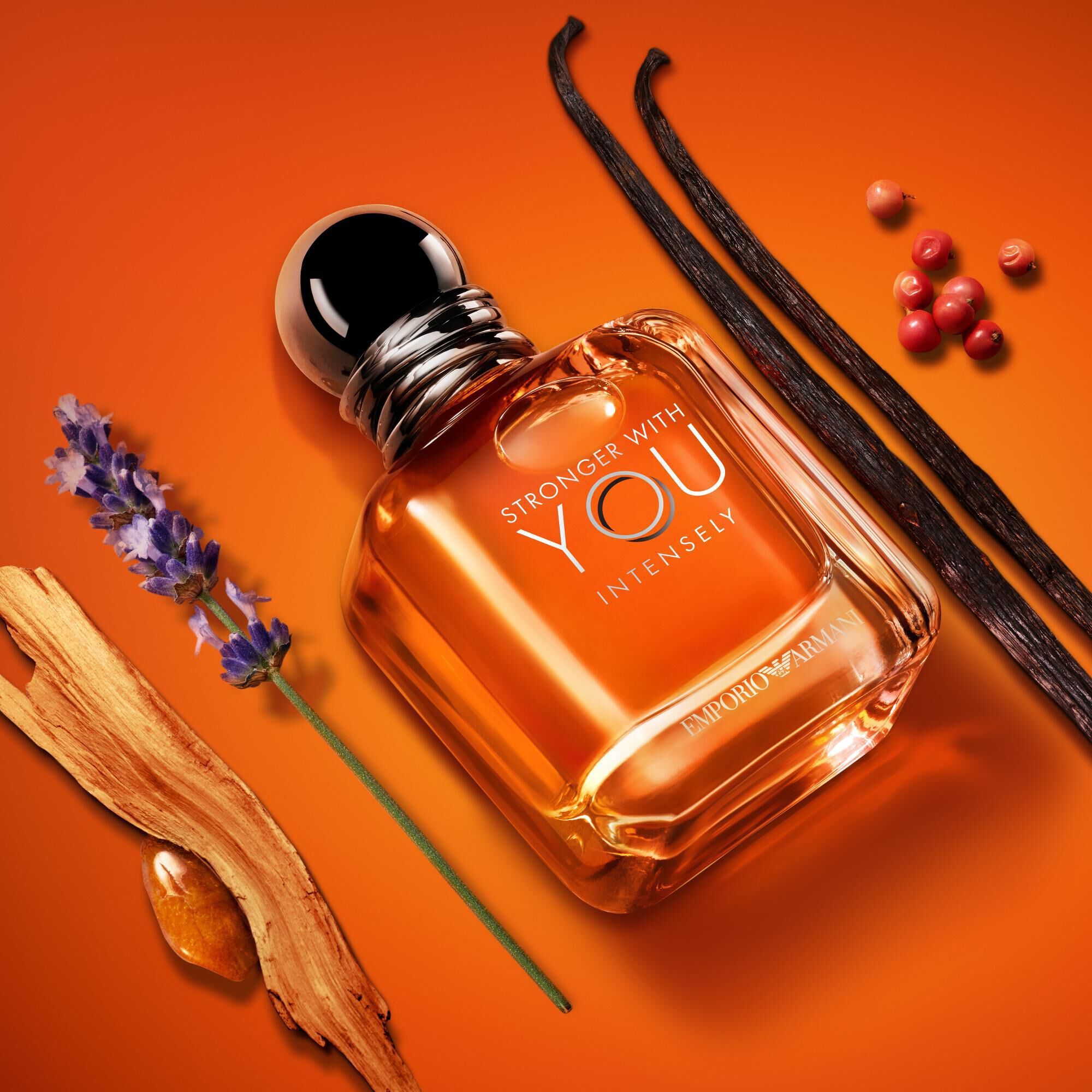 Emporio Armani Stronger With You Intensely Eau de Parfum — signature amber bottle surrounded by key olfactive ingredients: lavender, pink pepper, vanilla, and amber wood on a warm orange background.