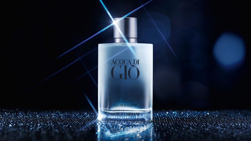 A bottle of Acqua di Gio fragrance with a clear, frosted appearance and a silver cap is centrally placed on a shimmering, reflective surface. A bright starburst light effect emanates from the top of the bottle, and the background is a dark, blurry blue with subtle light bokeh, creating a sophisticated and ethereal atmosphere.