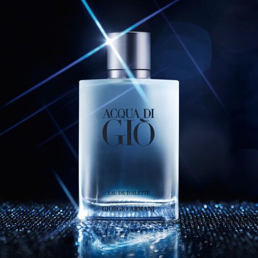 A bottle of Acqua di Gio fragrance with a clear, frosted appearance and a silver cap is centrally placed on a shimmering, reflective surface. A bright starburst light effect emanates from the top of the bottle, and the background is a dark, blurry blue with subtle light bokeh, creating a sophisticated and ethereal atmosphere.