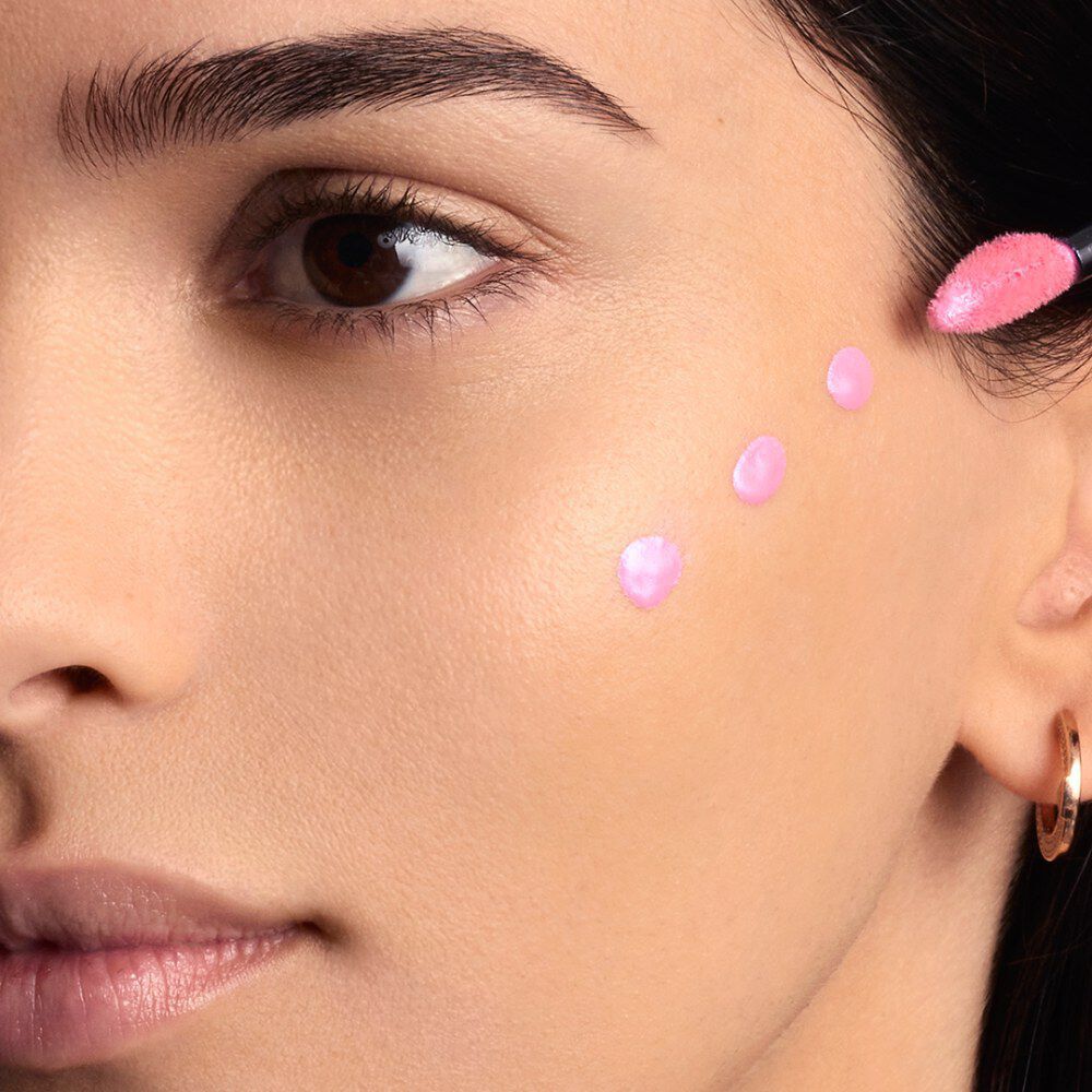 A woman's cheek is shown with five distinct dots of light pink liquid product, likely a blush or highlighter, dotted along her cheekbone area, ready for blending.