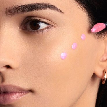 A woman's cheek is shown with five distinct dots of light pink liquid product, likely a blush or highlighter, dotted along her cheekbone area, ready for blending.