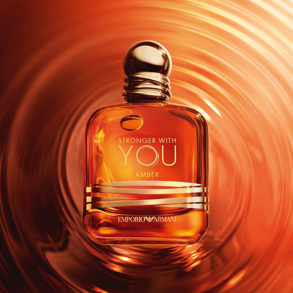 The Emporio Armani Stronger With You Amber fragrance bottle stands centered on a surface of rippling, warm amber-colored liquid, creating concentric circles around it.