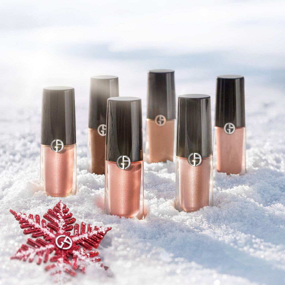 Five Armani beauty Eye Tint shades — from sheer rose to deep bronze — rest in fresh snow beside a crimson poinsettia. Holiday 2026 product flatlay.