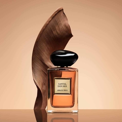 Armani Prive Les Eaux Santal Dān Shā eau de parfum, transparent rectangular glass bottle with black rounded cap, set against a warm terracotta and bronze sculptural background.