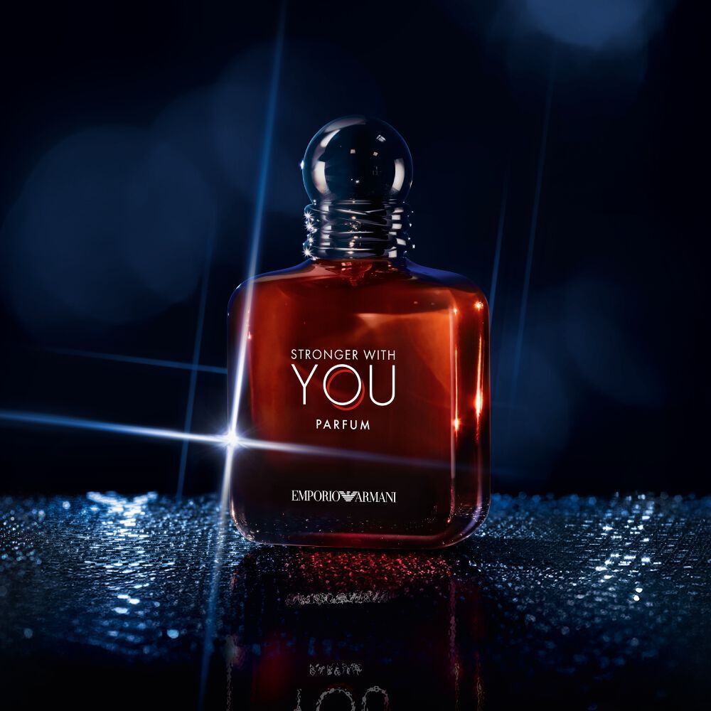  A single bottle of Emporio Armani "Stronger With You Parfum" stands upright on a reflective surface, illuminated by dramatic blue and white lighting against a dark background.