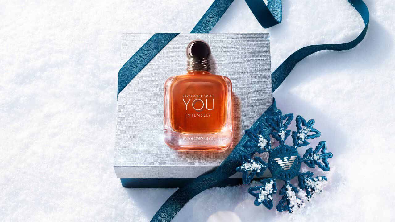 Emporio Armani Stronger With You Intensely Eau de Parfum — signature amber bottle resting on a silver Armani gift box with blue ribbon and ornamental snowflake, set against fresh white snow.