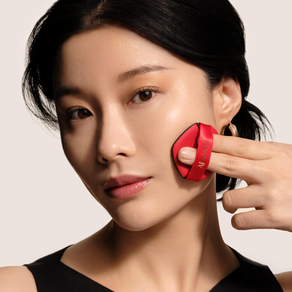 A model applying the LUMINOUS SILK PRIMA GLOW CUSHION to her cheek with the red Giorgio Armani cushion puff applicator, demonstrating the product in use.