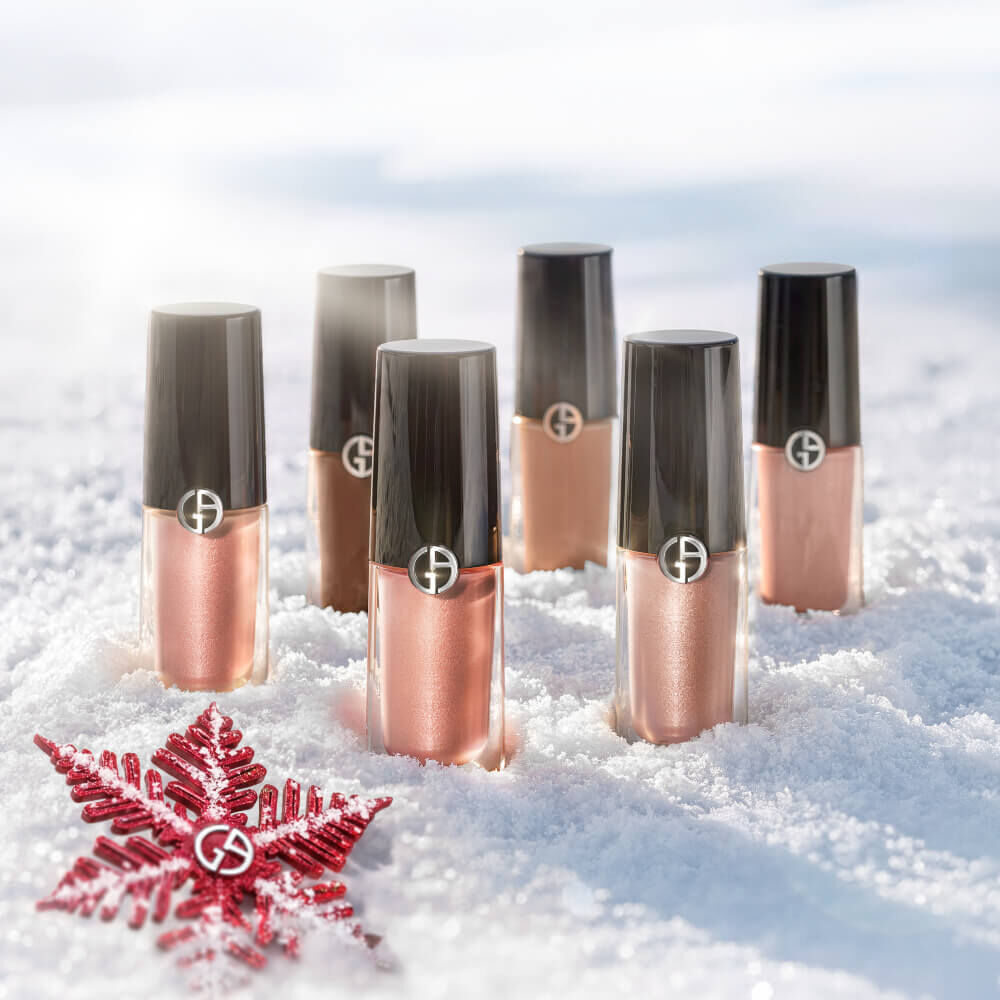 Five Armani beauty Eye Tint shades — from sheer rose to deep bronze — rest in fresh snow beside a crimson poinsettia. Holiday 2026 product flatlay.