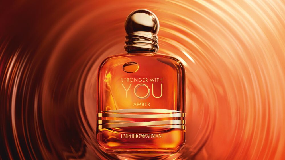 The Emporio Armani Stronger With You Amber fragrance bottle stands centered on a surface of rippling, warm amber-colored liquid, creating concentric circles around it.