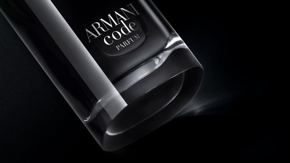 A close-up, angled view of the bottom of an Armani Code Parfum bottle, showcasing its clear glass base and part of the dark body with the silver logo, dramatically lit against a black background.