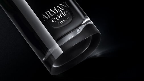 A close-up, angled view of the bottom of an Armani Code Parfum bottle, showcasing its clear glass base and part of the dark body with the silver logo, dramatically lit against a black background.
