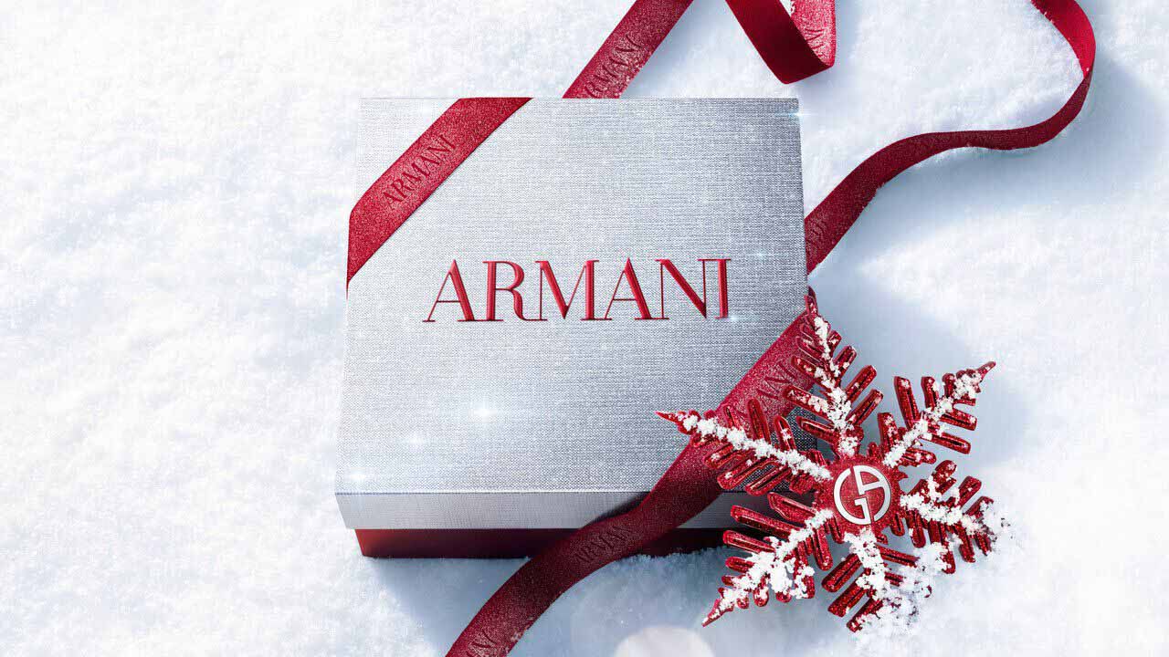 Silver Armani Glamourous Winter Holiday Gift Set box with red glitter ribbon and Giorgio Armani red snowflake ornament resting on a sparkling snowy surface — festive luxury fragrance gift packaging.