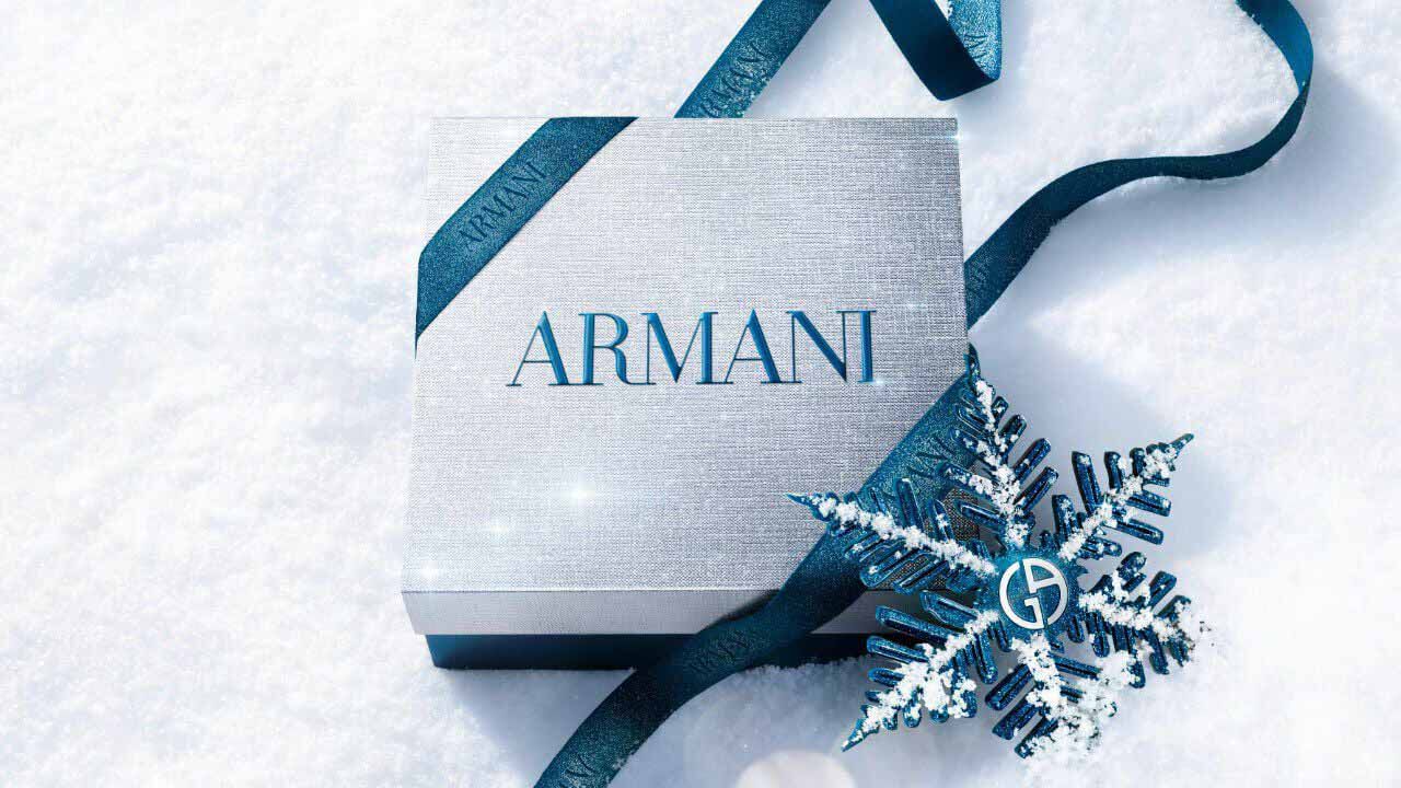 Armani beauty gift box — silver gift box wrapped with blue satin ribbon and a metallic snowflake ornament, resting on fresh white snow.