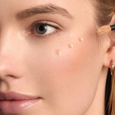 A woman's cheek is shown with five distinct dots of light pink liquid product, likely a blush or highlighter, dotted along her cheekbone area, ready for blending.