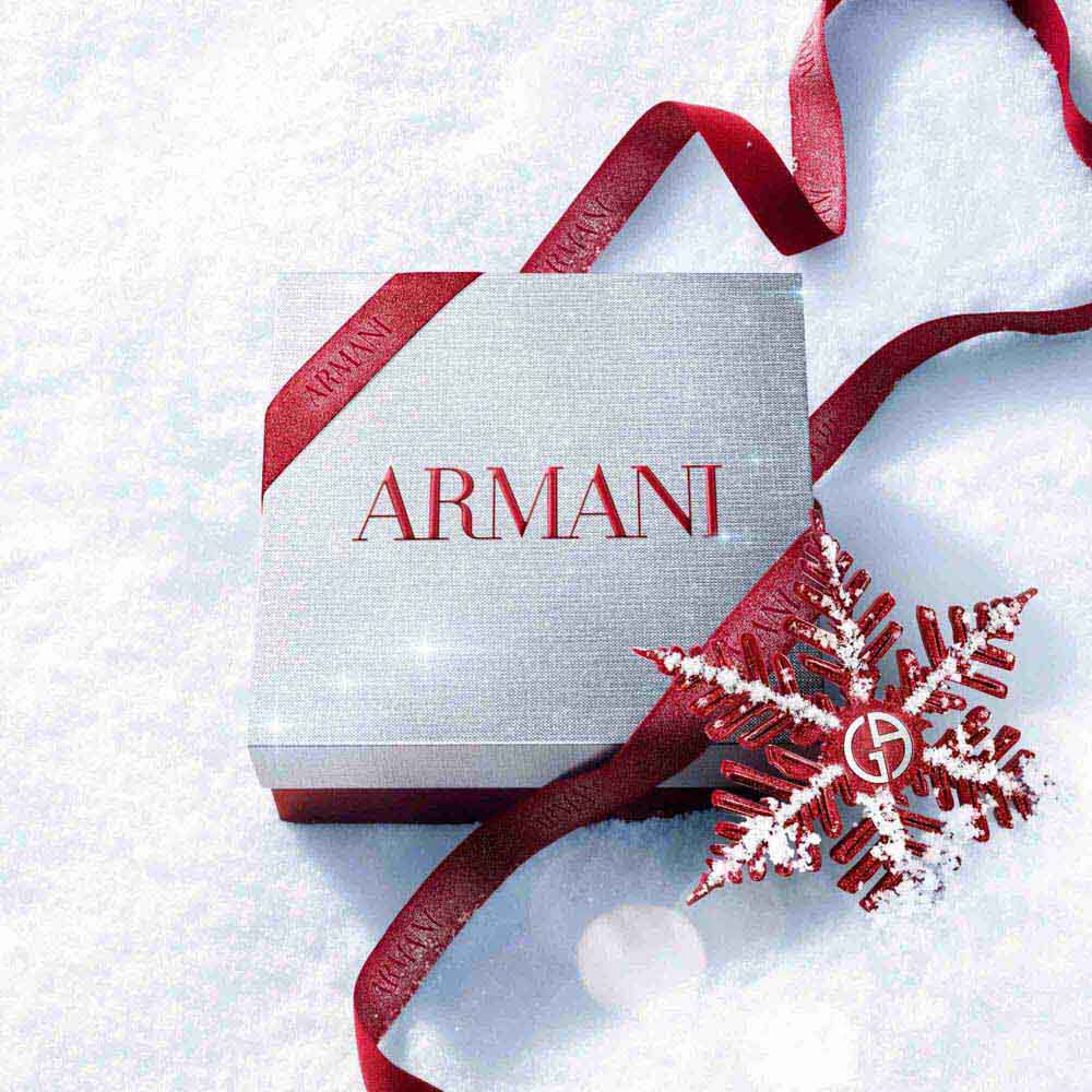 Silver Armani Glamourous Winter Holiday Gift Set box with red glitter ribbon and Giorgio Armani red snowflake ornament resting on a sparkling snowy surface — festive luxury fragrance gift packaging.