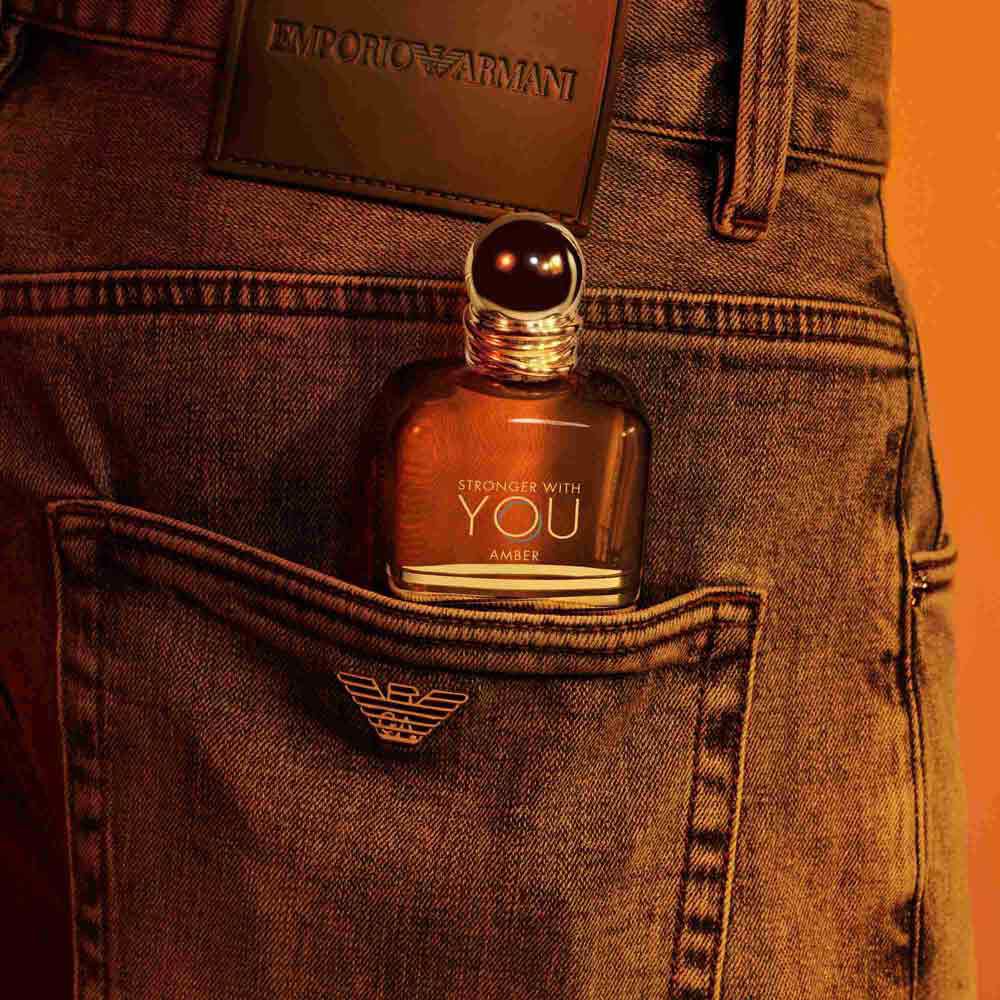 An Emporio Armani Stronger With You Amber fragrance bottle peeking out from the back pocket of a pair of jeans, with a dark leather patch visible above.