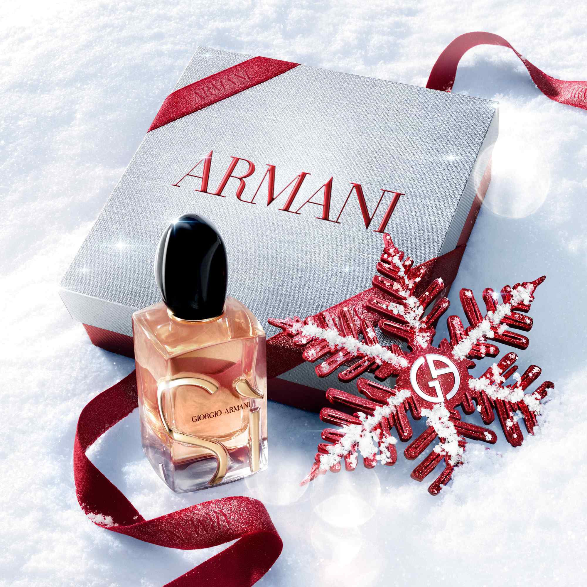 Si Eau de Parfum by Giorgio Armani, gold-toned bottle resting in snow with a red glitter ribbon and Giorgio Armani snowflake ornament — women's fragrance holiday