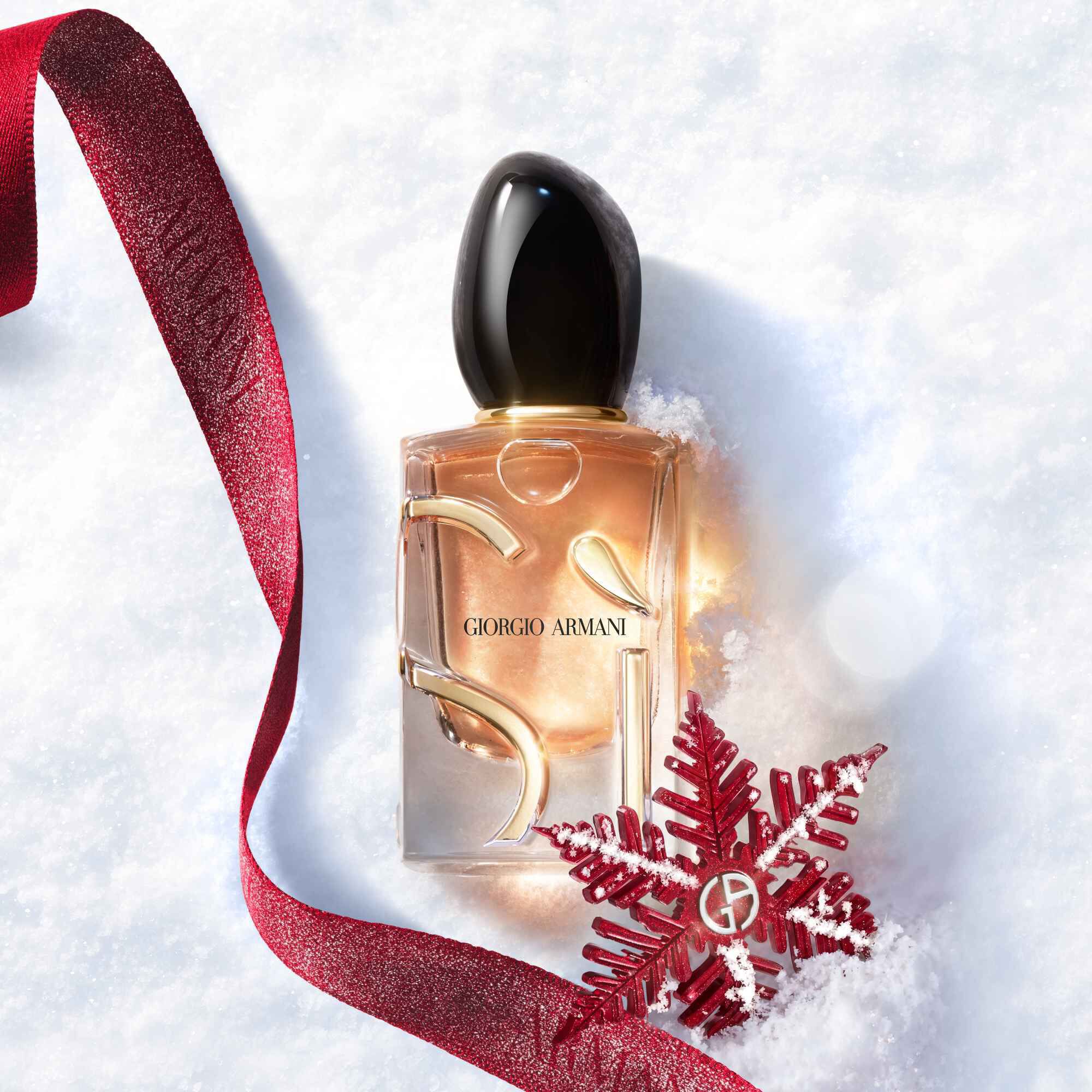 Si Eau de Parfum by Giorgio Armani, gold-toned bottle resting in snow with a red glitter ribbon and Giorgio Armani snowflake ornament — women's fragrance holiday gifting visual.