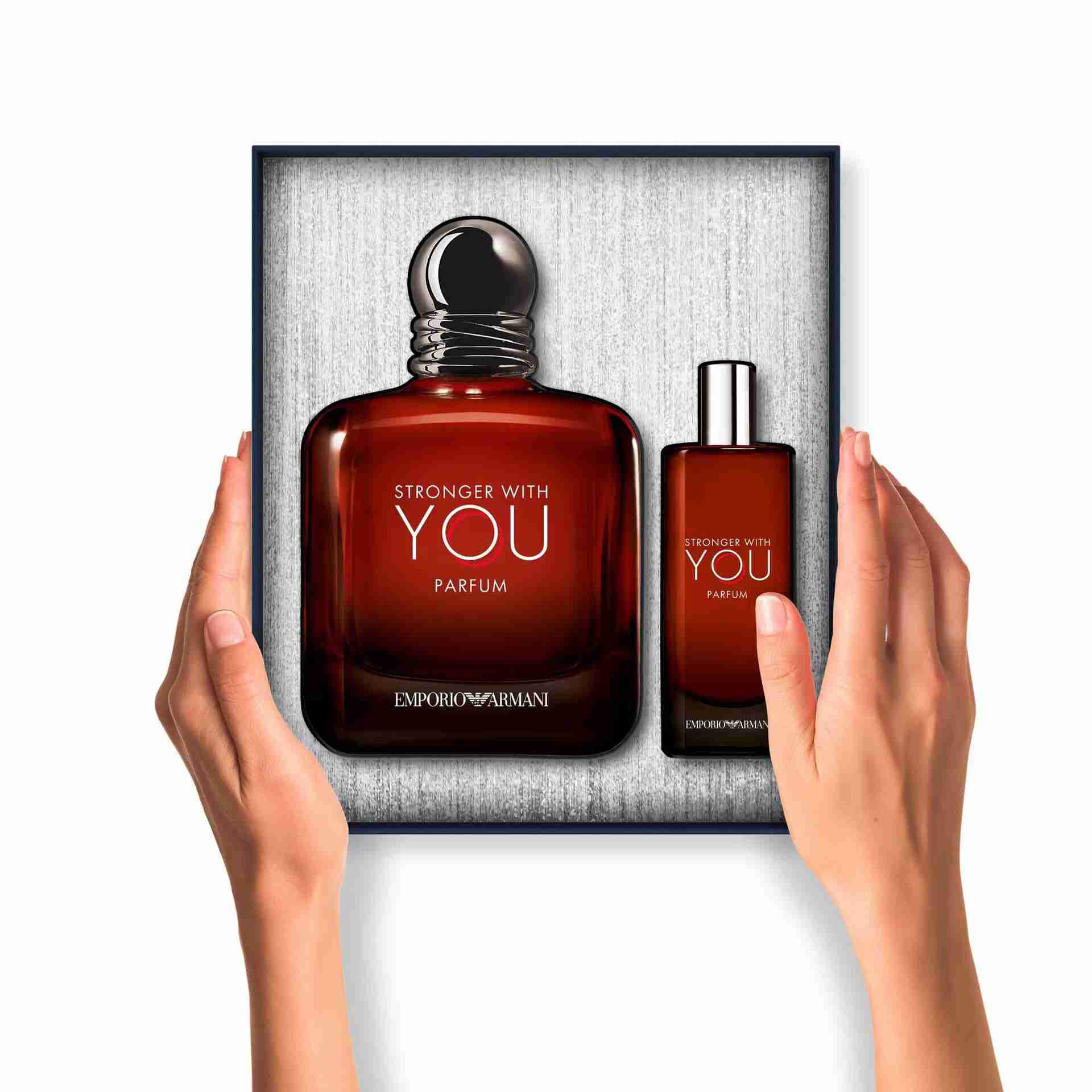 Two hands present an open gift box containing a large bottle and a travel-size spray of Emporio Armani "Stronger With You Parfum," both nestled inside the box.