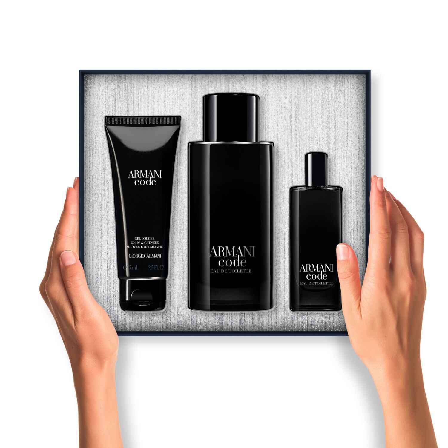 A pair of hands holds an open Armani Code gift box. Inside, three black-packaged products (body gel, large Eau de Toilette, and travel-size Eau de Toilette) are neatly arranged against a textured silver background.