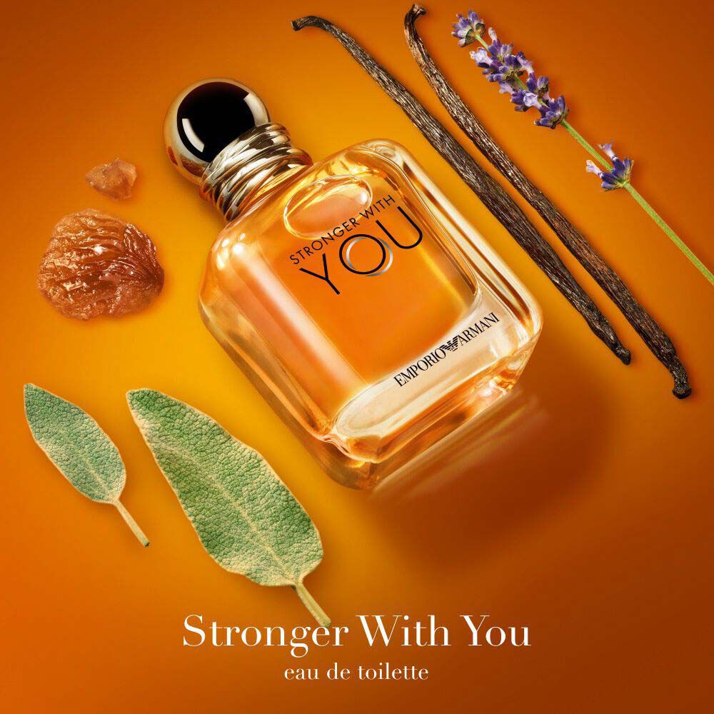 Emporio Armani Stronger With You Eau de Parfum — signature golden amber bottle surrounded by key olfactive ingredients: lavender, vanilla, chestnut and sage on a warm orange background.
