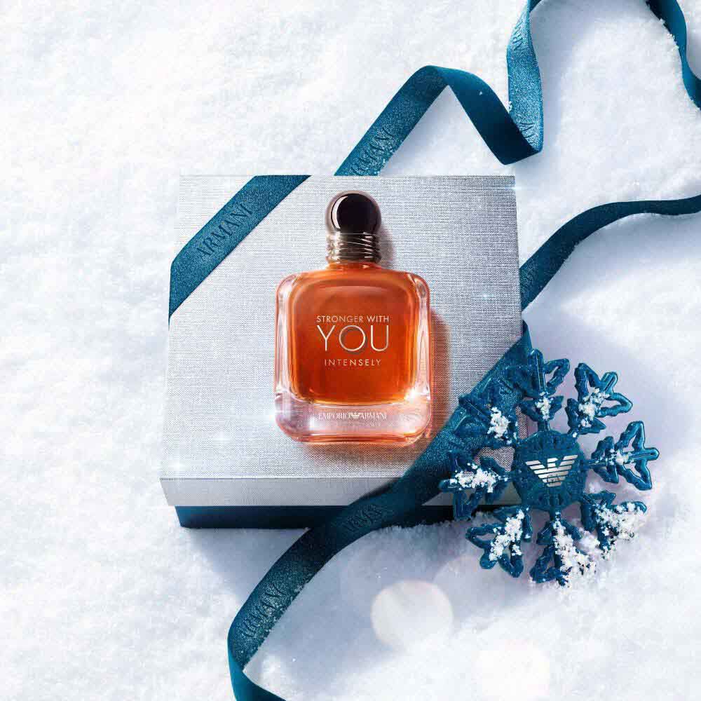 Emporio Armani Stronger With You Intensely Eau de Parfum — signature amber bottle resting on a silver Armani gift box with blue ribbon and ornamental snowflake, set against fresh white snow.