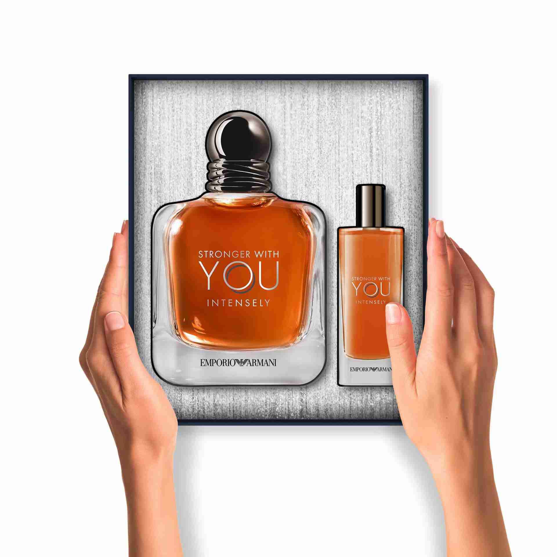 Two hands hold an open gift box containing a large bottle and a travel-size spray of "Stronger With You" Eau de Toilette, both neatly arranged inside the box.