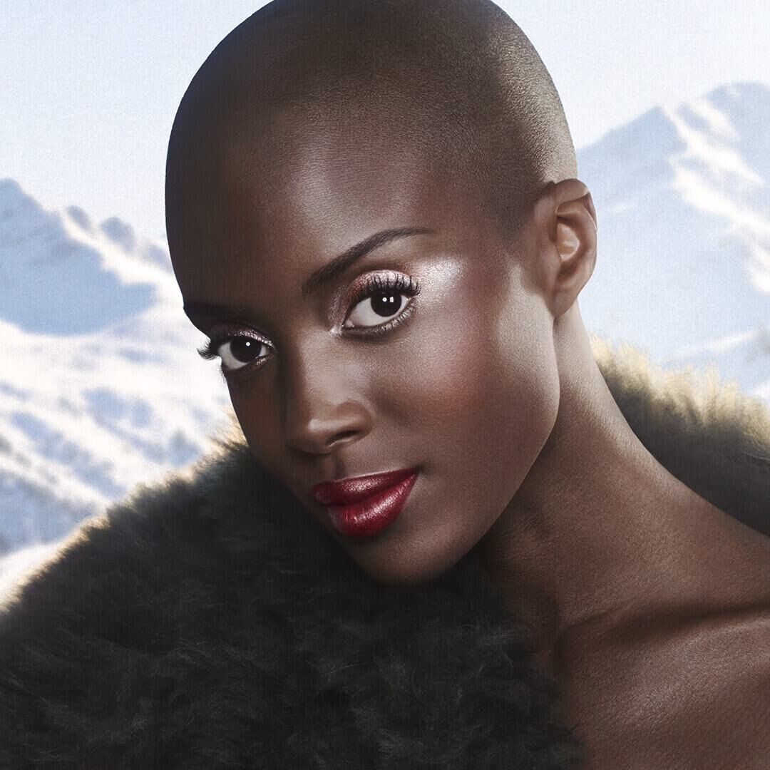 A model embodies winter elegance — Deep skin radiates an even, luminous glow. Eyes framed by full lashes and sculpted brows. A deep lacquered red lip as the defining statement. Snow-capped peaks behind. 