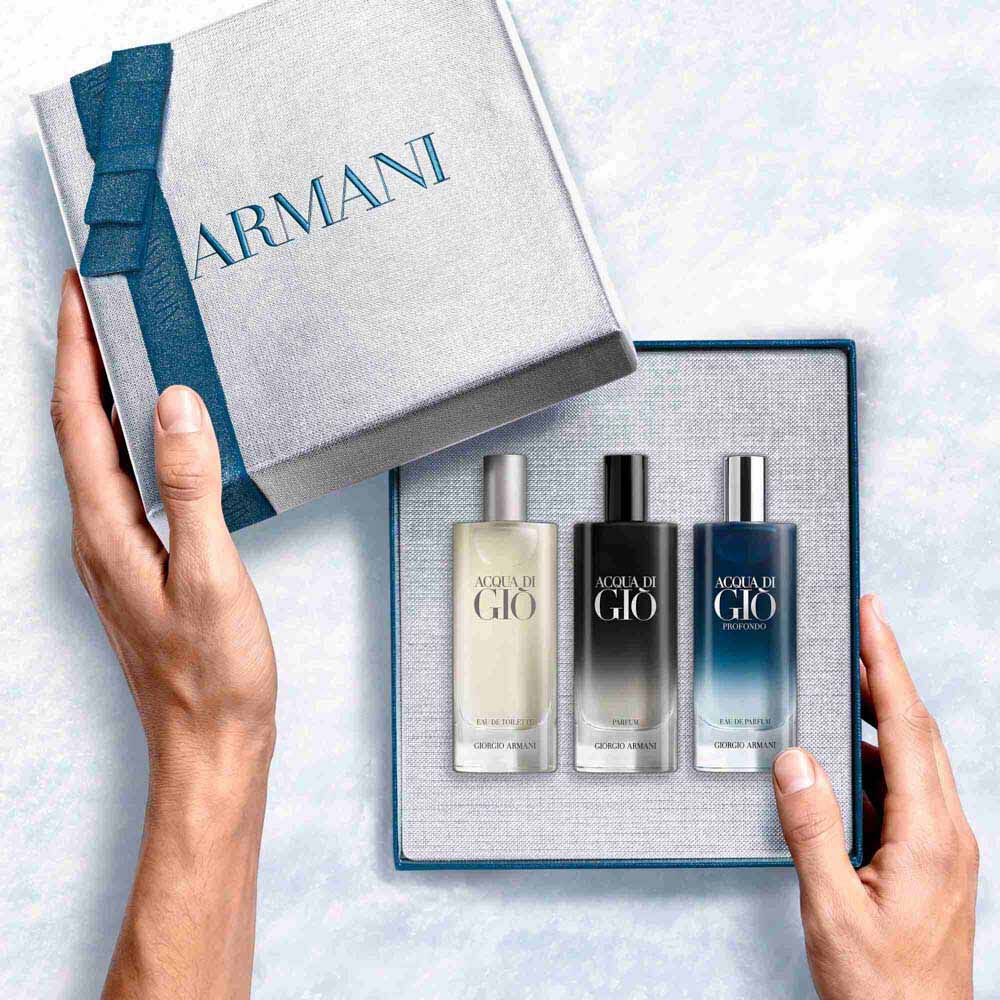 Hands opening a teal-ribboned Armani gift box revealing three Acqua di Gio mini bottles — Eau de Toilette, Profondo Eau de Parfum and Parfum 15ml discovery set.