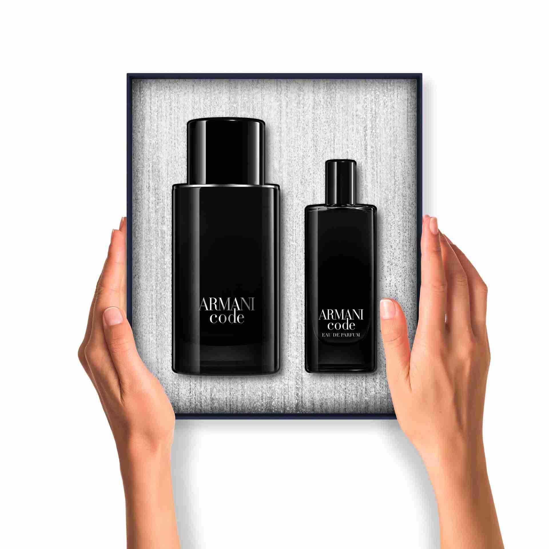 A pair of hands holds an open Armani Code gift box. Inside, two black-packaged products (body gel and Armani code Eau de Toilette) are neatly arranged against a textured silver background.
