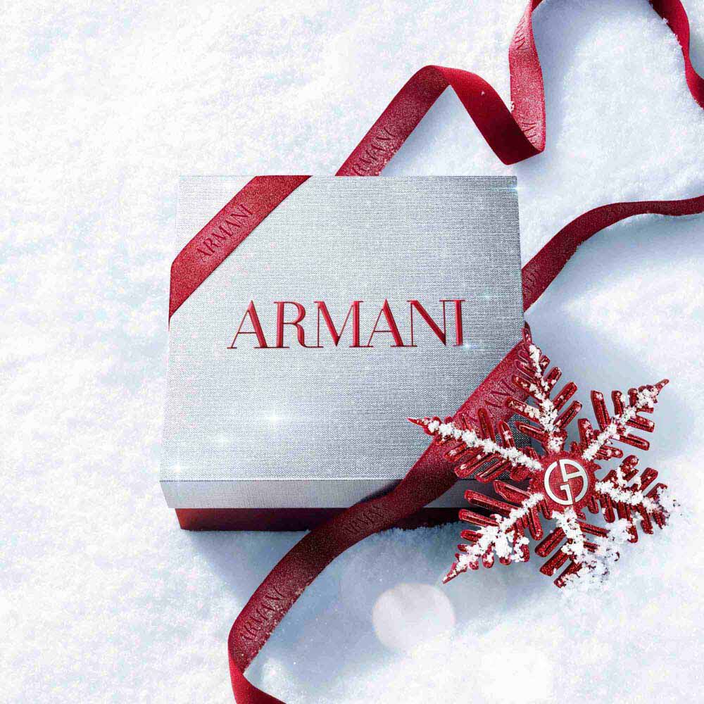 Silver Armani Glamourous Winter Holiday Gift Set box with red glitter ribbon and Giorgio Armani red snowflake ornament resting on a sparkling snowy surface — festive luxury fragrance gift packaging.