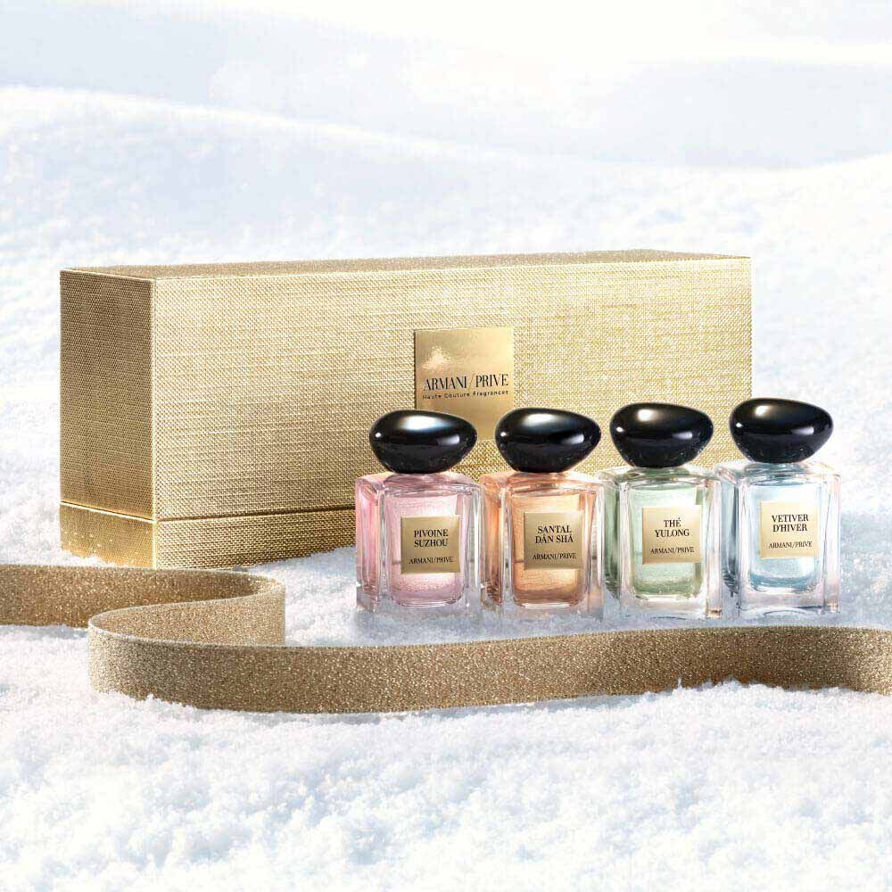 Armani Prive Les Eaux Holiday miniature gift set: gold rectangular box alongside four miniature eau de parfum bottles, photographed on a pale textured snow surface.