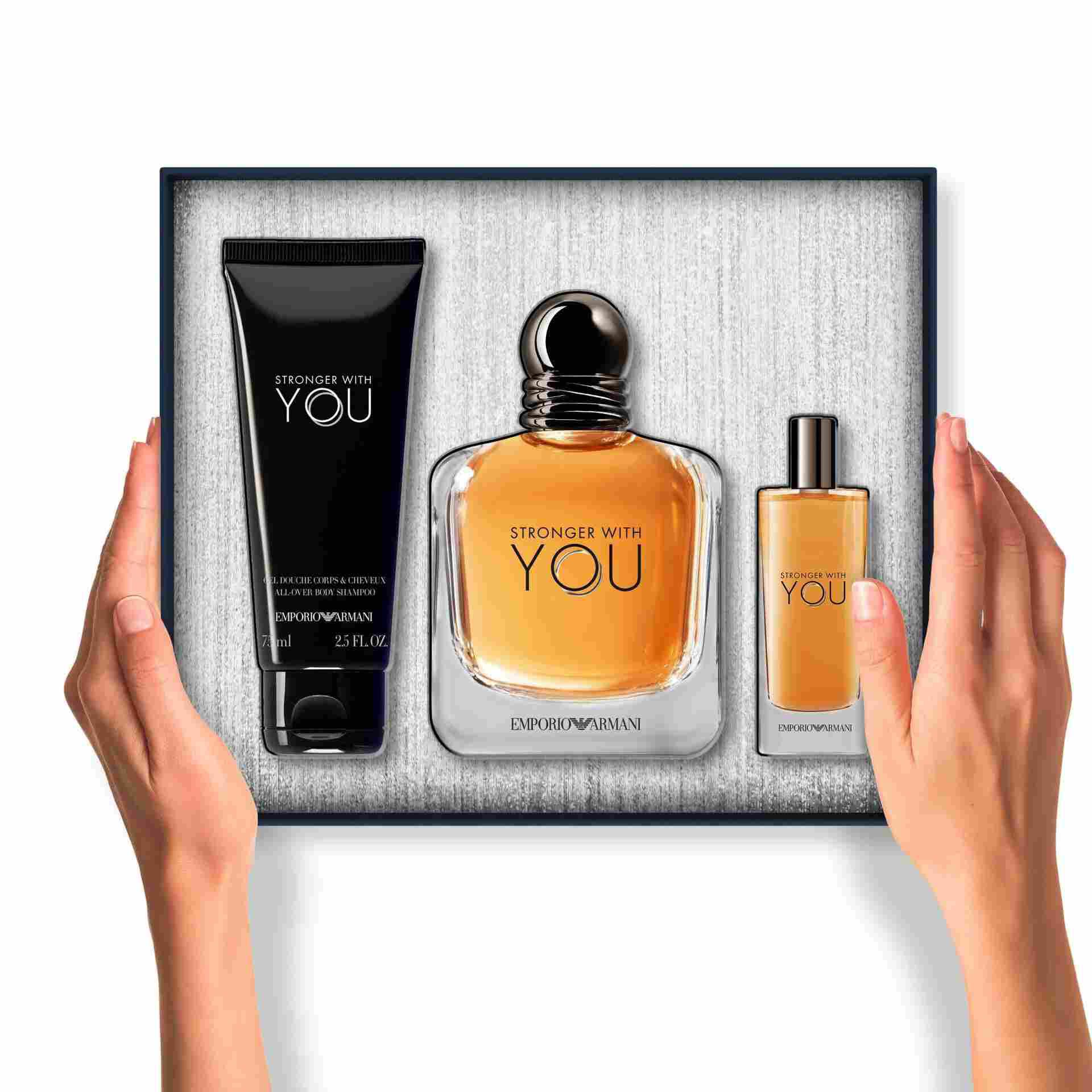 Two hands present an open gift box containing the Emporio Armani "Stronger With You" Eau de Toilette, a travel-size spray, and an All-Over Body Shampoo, all neatly arranged inside the box.