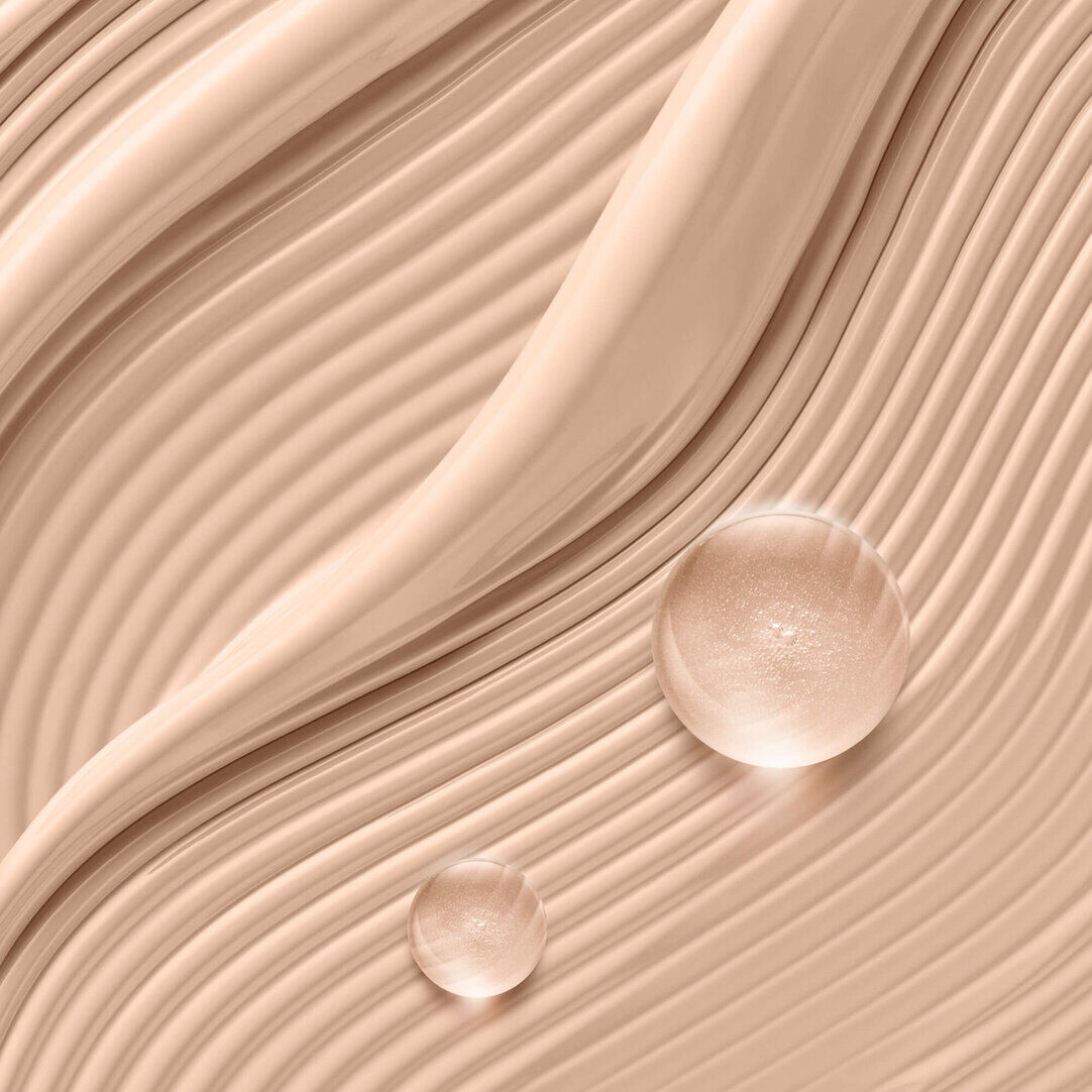 Close-up of a smooth, wavy foundation texture in a light beige shade, with two clear, luminous spheres showcasing the product's hydrating properties.