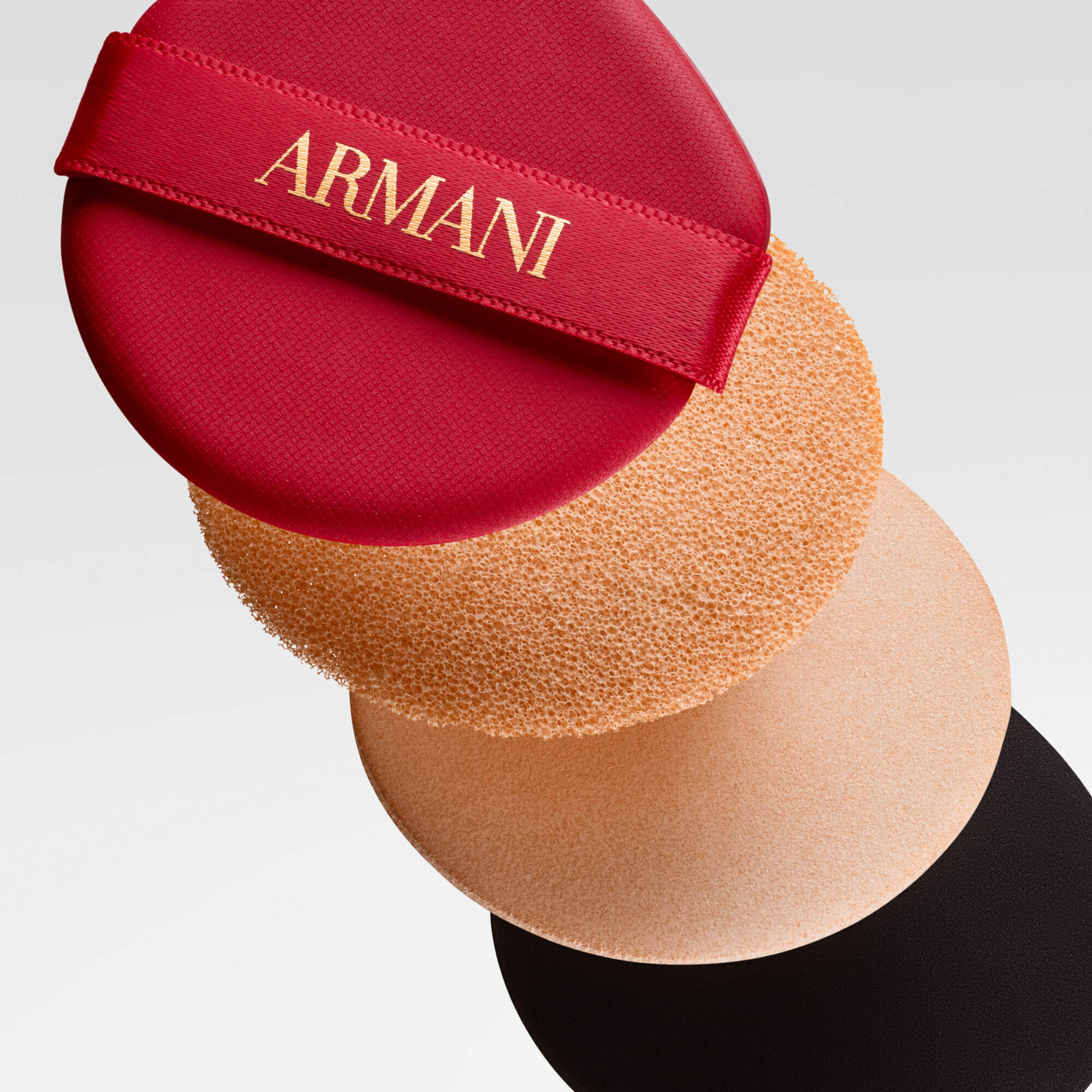 A stack of four round cushion foundation elements, including a red applicator puff with ARMANI logo in gold, a beige cushion, a lighter beige sponge, and a dark base, showcasing the product's layers.