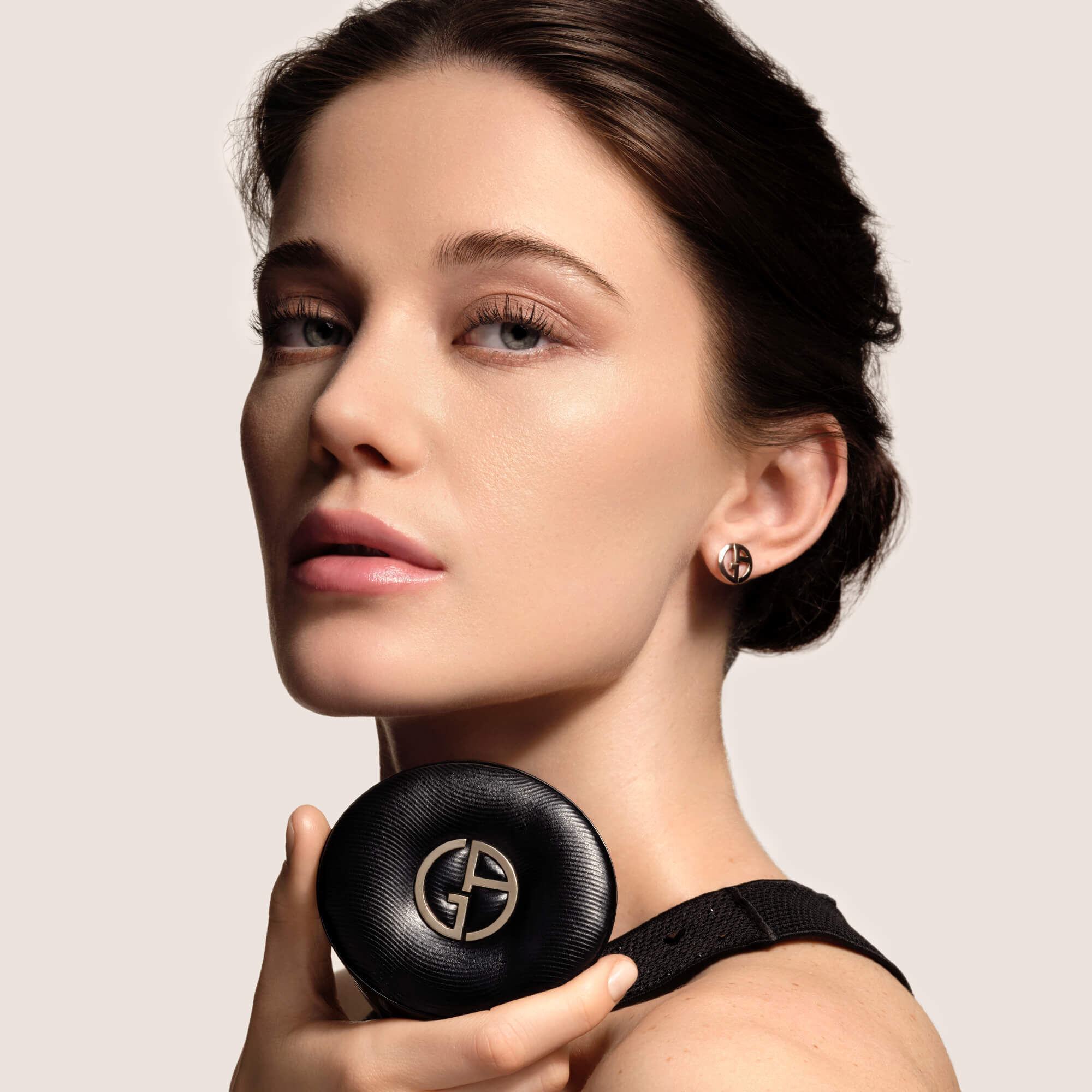 A model with elegant hair and natural makeup holds the black Giorgio Armani LUMINOUS SILK PRISMA GLOW CUSHION compact, showing the product in use.