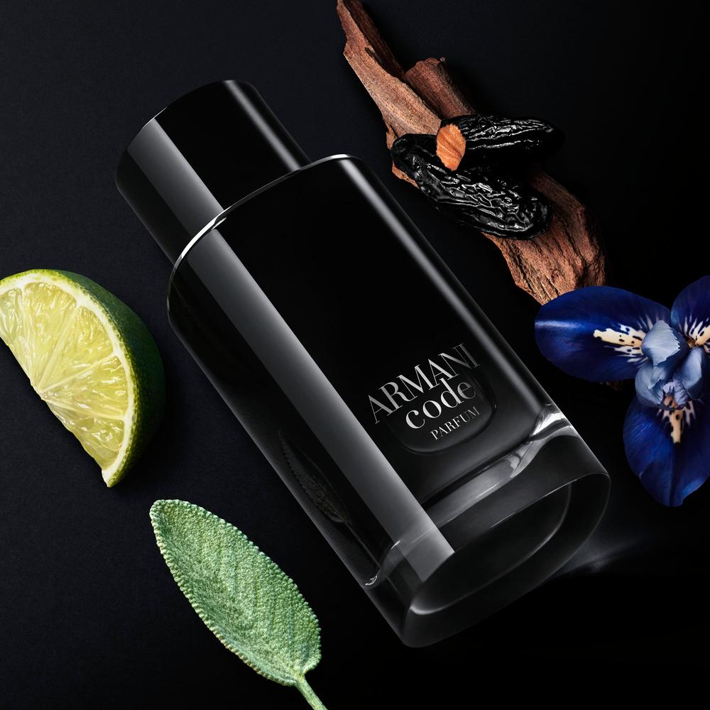 A black Armani Code parfum bottle with a silver label lies on textured black leather, next to a slice of green mandarin and grilled tonka beans, highlighting the fragrance’s fresh and warm key ingredients.