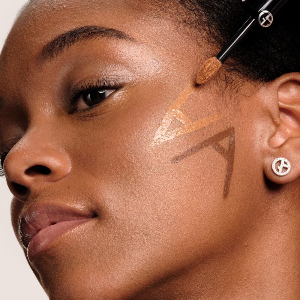 Close-up of a model with deep skin having Luminous Silk Concealer applied to her cheek in a geometric line pattern using the wand applicator.