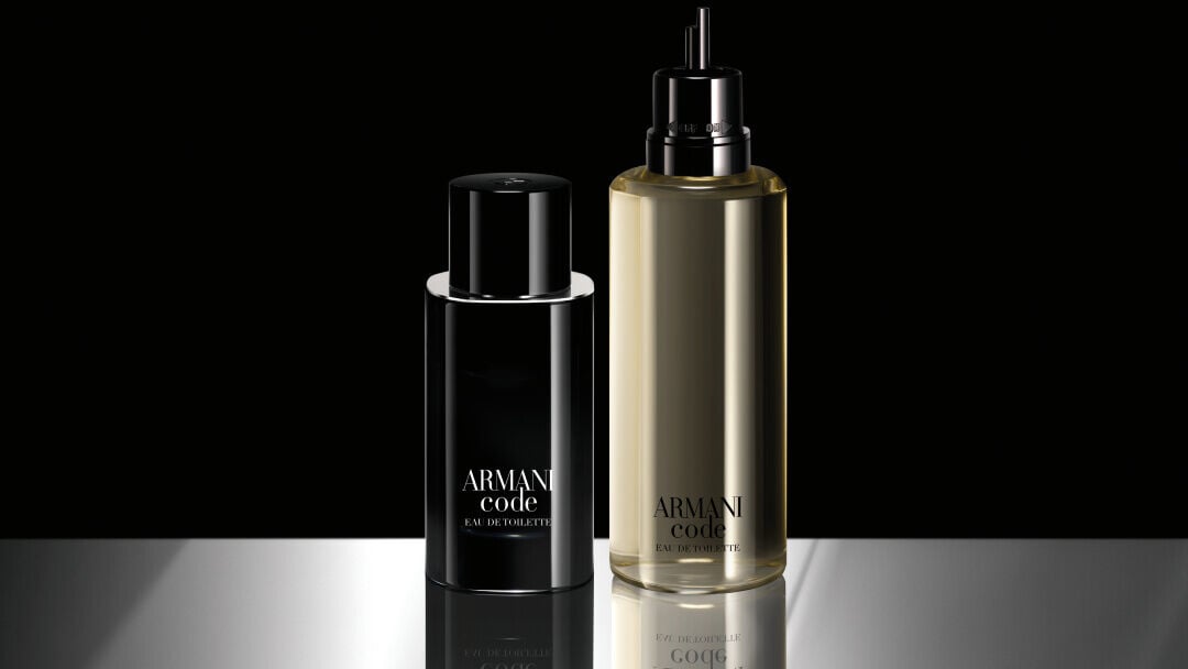 Side-by-side display of a full ARMANI CODE EAU DE TOILETTE bottle and a larger refill bottle. The refill bottle is transparent, revealing the pale yellow fragrance inside, and both have the Armani Code branding in white.